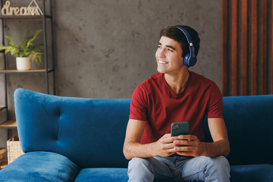 Young smiling man wears red t-shirt headphones listen music hold use mobile cell phone sit on blue sofa couch stay at home hotel flat rest relax spend free spare time in living room indoors grey wall.