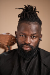 Fototapeta premium A close-up portrait of an adult Black man with dreadlocks and a beard wearing wireless earbuds as a barber trims his hair with clippers during a grooming session in a studio.