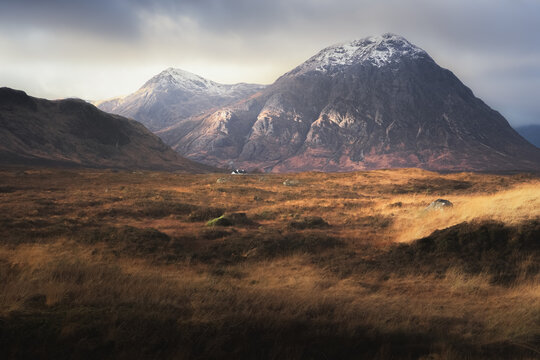 Dramatic golden light on the dark, moody, mountain landscape of Buachaille Etive Mor with a distant Blackrock Cottage at Glencoe in the Scottish Highlands, Scotland.