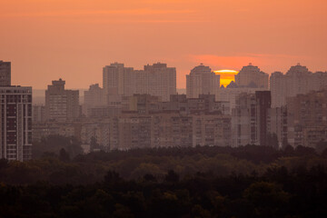 Sun rising over residential high rise buildings and Dnipro river. Urban cityscape at dawn with orange sky, morning haze and atmospheric city landscape silhouette in golden sunrise light.