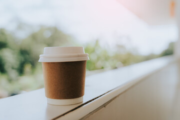 A coffee cup sits on a balcony railing. The cup is brown with a white lid. The blurred background reveals natural light and the green of the trees, creating a simple and relaxing atmosphere, perfect f
