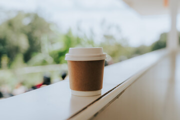 A coffee cup sits on a balcony railing. The cup is brown with a white lid. The blurred background reveals natural light and the green of the trees, creating a simple and relaxing atmosphere, perfect f