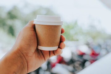 A coffee cup sits on a balcony railing. The cup is brown with a white lid. The blurred background reveals natural light and the green of the trees, creating a simple and relaxing atmosphere, perfect f