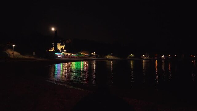 The full moon and colorful night lights of a beach bar reflecting on the calm sea surface in Njivice, Croatia. Njivice is a coastal village located on Krk Island in Primorje-Gorski Kotar County.
