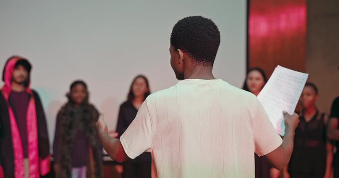 African young adult gen z male man hold scripts paper read play story lines scene standing indoor stage hall. Happy group boy girl do work drama practice day studio campus place