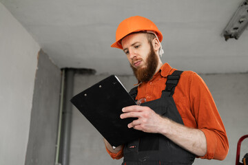 Young handyman man wear helmet hold clipboard with documents standing in new apartment. renovation...