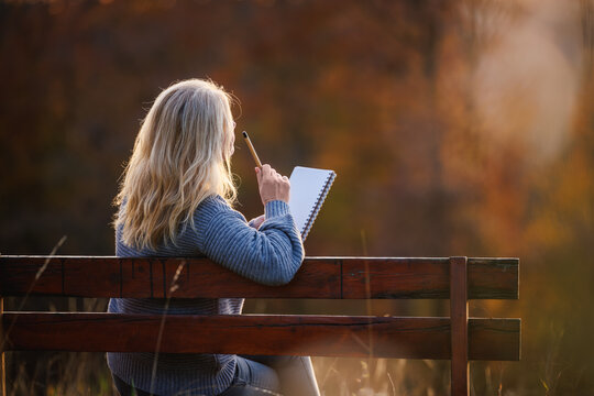 Thoughtful woman sitting on park bench and writing diary or inspiration notes during sunset. Journaling outdoors for self improvement