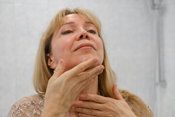 Woman in her 50s performing facial massage with handheld skincare tool in bathroom, wellness and anti-aging theme. Female self-care concept