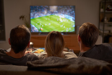 Three watching a football match on tv on a couch at home, view from back
