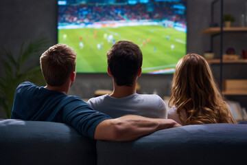 Three watching a football match on tv on a couch at home, view from back