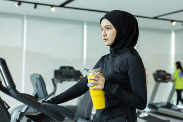 Muslim women in sportswear holding yellow water bottle while standing on treadmill at gym. Promotes hydration, fitness, wellness lifestyle. Ideal for health ads, hijab fitness and gym visuals.