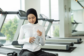 Muslim woman in white sportswear sitting on treadmill and holding water bottle after workout. Reflects rest, hydration, healthy lifestyle. Ideal for fitness, wellness and hijab sports themes.