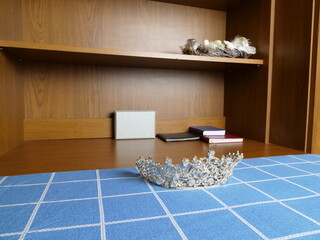 Decorative silver crown with stones lying on a blue checkered surface against shelves background.