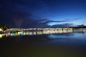 The historic Truong Tien Bridge radiates purple light across the river at night, hovering above a passing traditional dragon boat