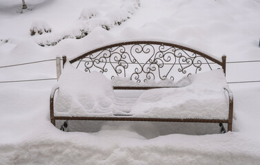 Snow-covered park bench during winter snowfall, symbolizing cold weather, seasonal stillness and quiet urban outdoor space with empty seating and soft snow textures.