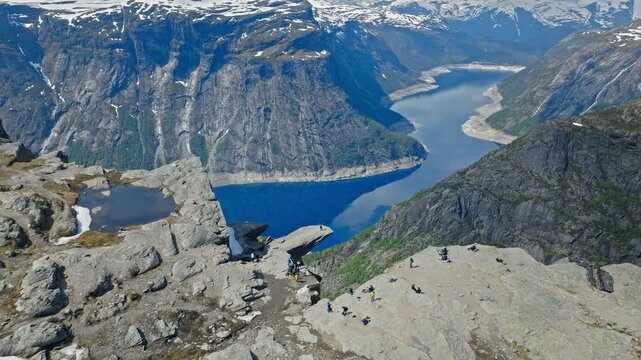 Rocky mountain slope descending toward the fjord at Trolltunga. Aerial summer view combining rugged stones, snow traces, and vivid blue water below.