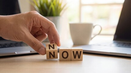 Adult mans hand arranging wooden blocks spelling NOW on a desk with laptops and coffee cup, symbolizing urgency and present moment