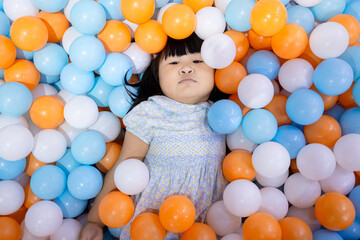 Adorable Little Asian Girl Relaxing and Lying in Colorful Plastic Ball Pit,A cute young Asian girl lying down and covered with many blue, orange, and white plastic balls in a playground ball pit,