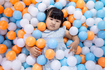 Adorable Little Asian Girl Relaxing and Lying in Colorful Plastic Ball Pit,A cute young Asian girl lying down and covered with many blue, orange, and white plastic balls in a playground ball pit,