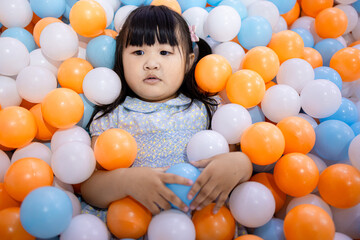 Adorable Little Asian Girl Relaxing and Lying in Colorful Plastic Ball Pit,A cute young Asian girl lying down and covered with many blue, orange, and white plastic balls in a playground ball pit,