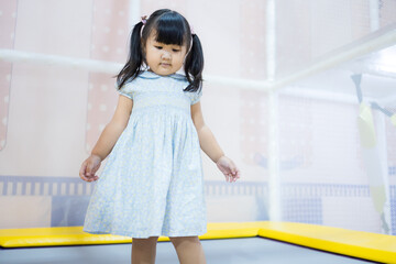Cute Little Asian Girl Jumping and Playing on Trampoline in Indoor Playground,A cheerful young Asian girl in a blue dress enjoying her time jumping on a trampoline at an indoor soft play center.
