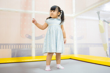 Cute Little Asian Girl Jumping and Playing on Trampoline in Indoor Playground,A cheerful young Asian girl in a blue dress enjoying her time jumping on a trampoline at an indoor soft play center.