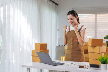 Asian woman feeling happy and successful, celebrating a new order or good sales from her online business, packing products at home