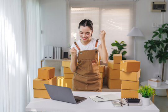 Young woman entrepreneur celebrating her business growth with numerous shipping boxes, a laptop, and a notebook on a desk