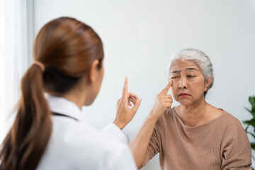 Asian doctor examining senior woman's eye, pointing a finger for eye movement test. Patient feels...