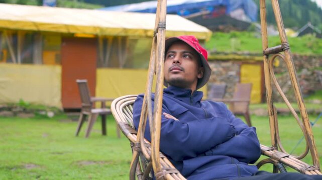An adult Indian man happily playing on a swing during leisure time, enjoying and eating an apple in Kheerganga, Kasol, Himachal Pradesh, India.