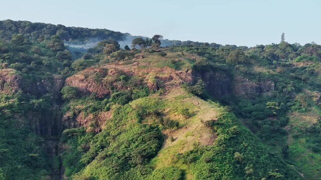 Side view of Sahyadri mountain cliffs with green trees, dry grass, rocky textures and natural terrain in daylight. Lush forest meets steep rugged rock faces.