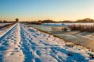 Dutch polder landscape in winter. A snow-covered footpath on a dike is between the fields and ditches. The photo was taken at the end of a winter day with the sun already low in the sky.