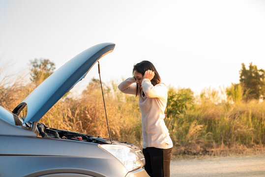 A woman showing frustration and stress by holding her head while looking at the open hood of her broken-down car on a quiet road during sunset.