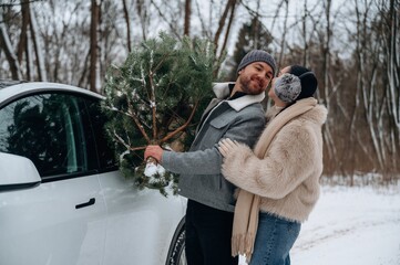 Couple joyfully carrying a Christmas tree from a car in a snowy landscape, celebrating the holiday...