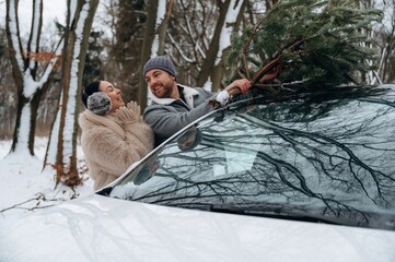 Couple joyfully securing a Christmas tree on top of their car in a snowy forest, showcasing festive...