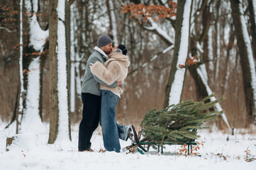 Couple embracing in snowy forest, surrounded by trees, with a sled carrying a freshly cut Christmas tree, capturing a joyful winter moment of love and celebration