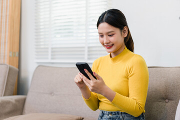 Smiling Asian woman using smartphone and social media app while sitting on a sofa in a bright living room