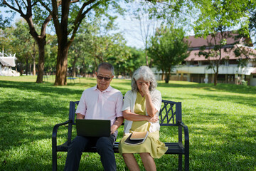 Elderly Asian couple sitting on a park bench as the man uses a laptop while the woman appears stressed and worried.
