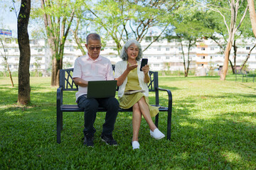 Senior Asian couple using a laptop and smartphone while sitting together on a park bench in a sunny outdoor setting