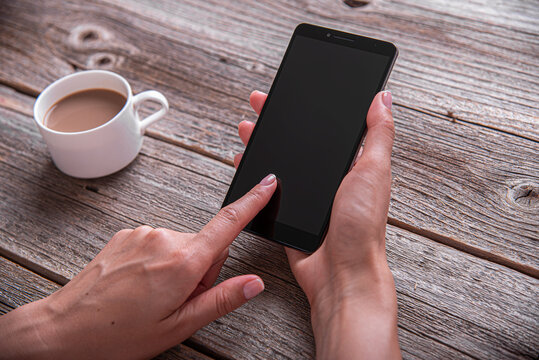 A person holding a black smartphone over a rustic wooden table next to a cup of coffee.