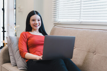 Happy young Asian woman using a laptop while sitting on a sofa in a bright living room.