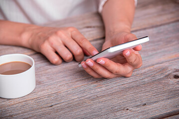 A woman uses a white smartphone while sitting at a rustic wooden table next to a cup of coffee.