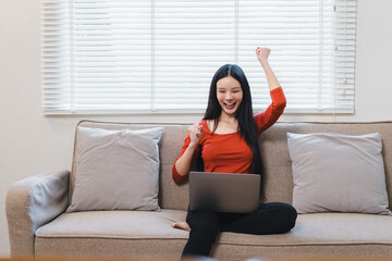Happy young Asian woman celebrating success while sitting on a sofa with a laptop in a bright living room