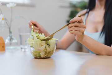 A young woman enjoying a fresh green salad with a gold fork from a glass bowl, focusing on a healthy lifestyle and nutrition.