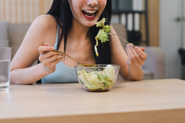 Happy young Asian woman eating fresh green salad with golden fork and spoon at home for a healthy lifestyle