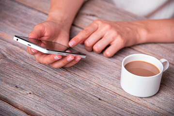 A woman uses a white smartphone while sitting at a rustic wooden table next to a cup of coffee.
