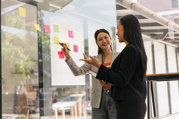 Fototapeta premium Two professional Asian businesswomen brainstorming and discussing project ideas with colorful sticky notes on a glass wall.