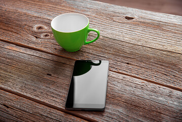 A black smartphone and an empty green ceramic mug sitting on a rustic wooden table surface.
