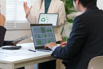 A professional businessman uses a laptop to analyze ESG data while a colleague presents sustainable business metrics on a digital tablet in a bright office meeting.