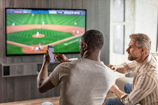 Two friends holding smartphone with sports bets app while watching baseball match at home in living room